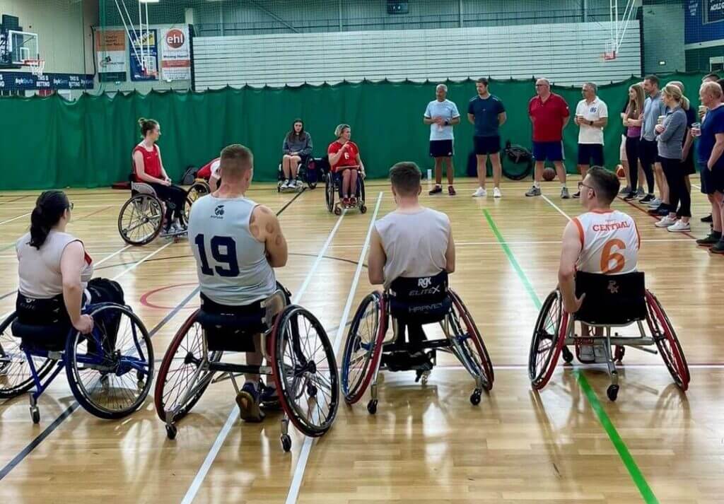 A group of wheelchair basketball players in a gym, wearing athletic gear, listen to a coach. Spectators stand nearby. The mood is focused and attentive.