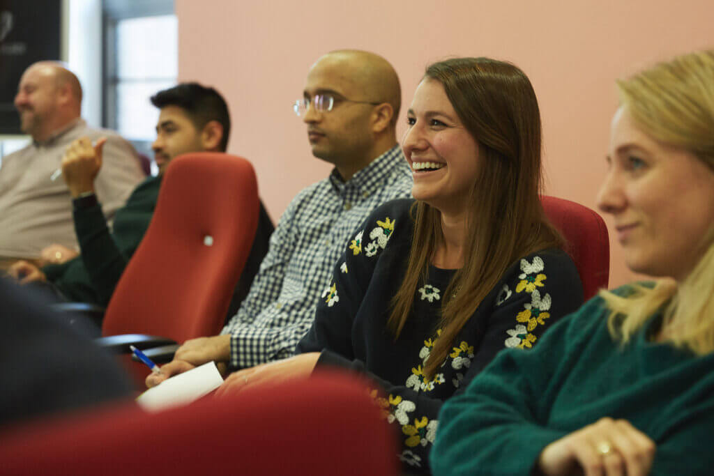 A group of people seated in red chairs faces forward, attentively engaged and smiling during a meeting or presentation in a warmly lit room with pink walls.
