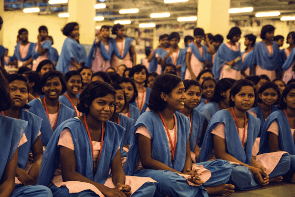 A large group of smiling young women are seated and standing in a brightly lit indoor setting. They wear matching blue uniforms with pink tops, creating a sense of unity and camaraderie. The mood is cheerful and lively.