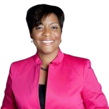 A woman with short dark hair is smiling and wearing a bright pink blazer over a black top, standing against a plain white background.
